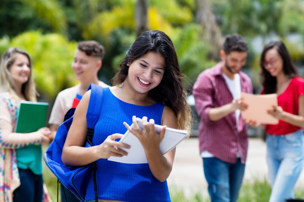 Young female Hispanic student with backpack smiles down at the papers in her hand while walking across a college campus, with blurred students in the background