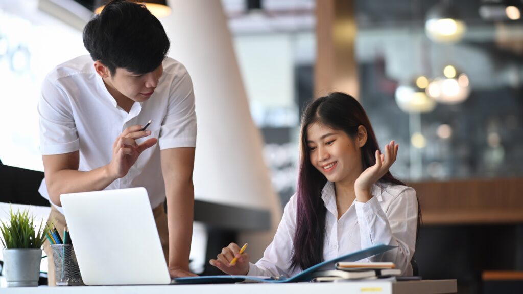 Young female Asian MBA student, wearing a white shirt, sitting with books and a laptop. Her male mentor stands beside her, wearing a white dress shirt.