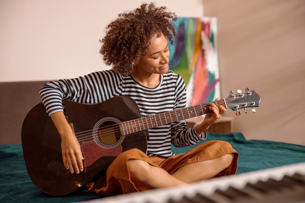 Multiracial, minority young woman playing guitar at her home. An out of focus keyboard is in front of her.
