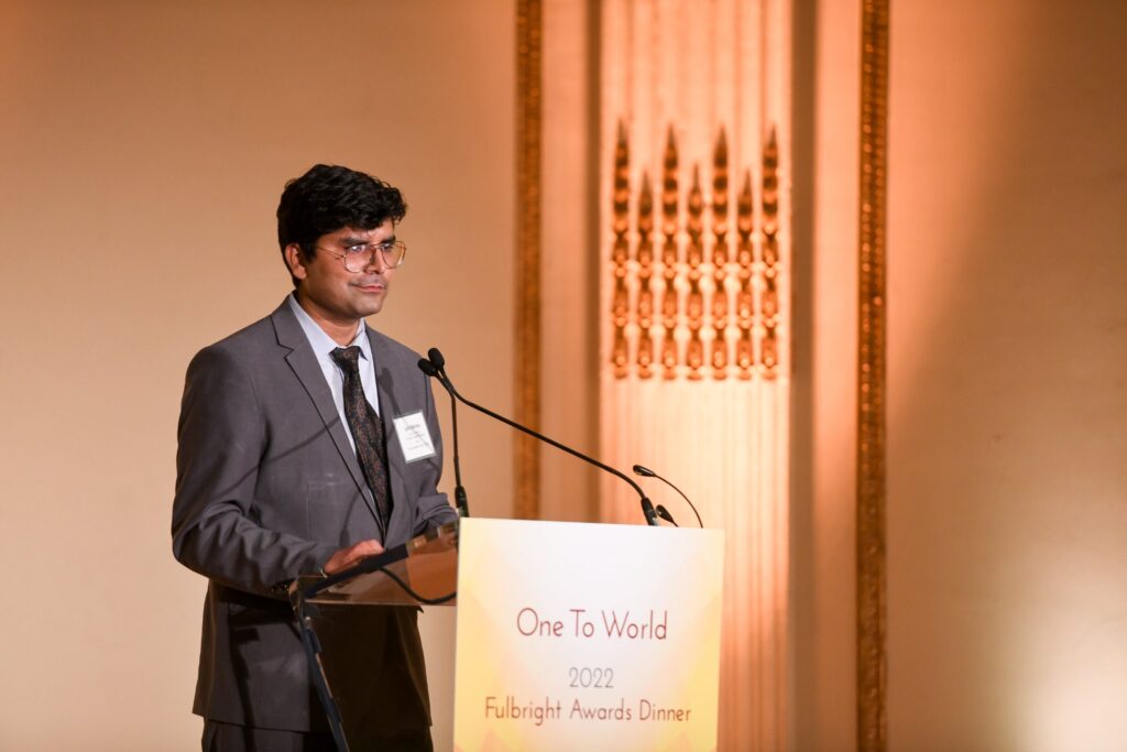 Fulbright Nehru scholar Dr. Moham stands at a podium delivering his speech. The podium has a banner that reads "One to World 2022, Fulbright Awards Dinner."