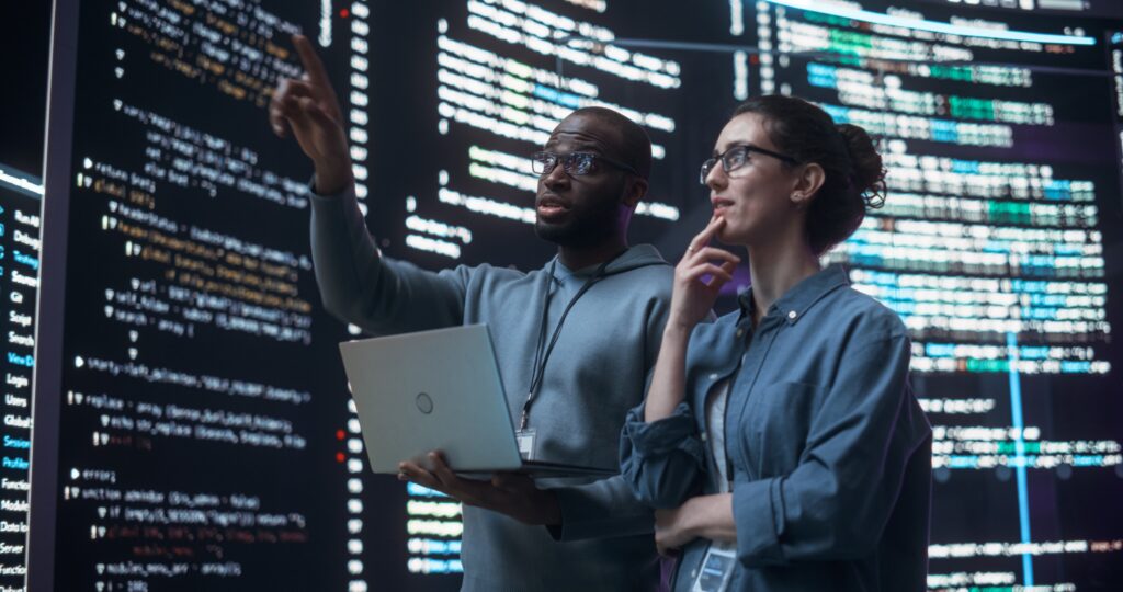 A black man and an ethnically ambiguous woman stand in front of wall-sized tech screens with lines of code on them, discussing with a laptop in their hands.