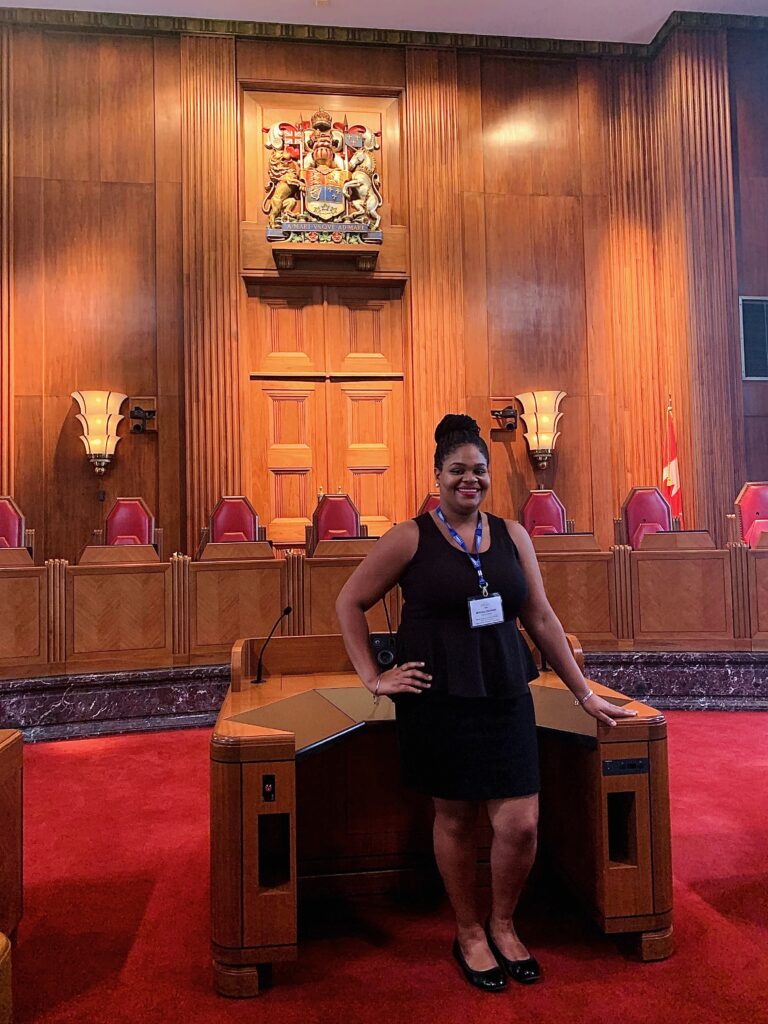 Brittany inside the Supreme Court of Canada during the Fulbright Killam Fellowship. She stands in front of a large, wooden speaking podium. Behind her are raised chairs and a fully wooden paneled wall with a crest near the ceiling.