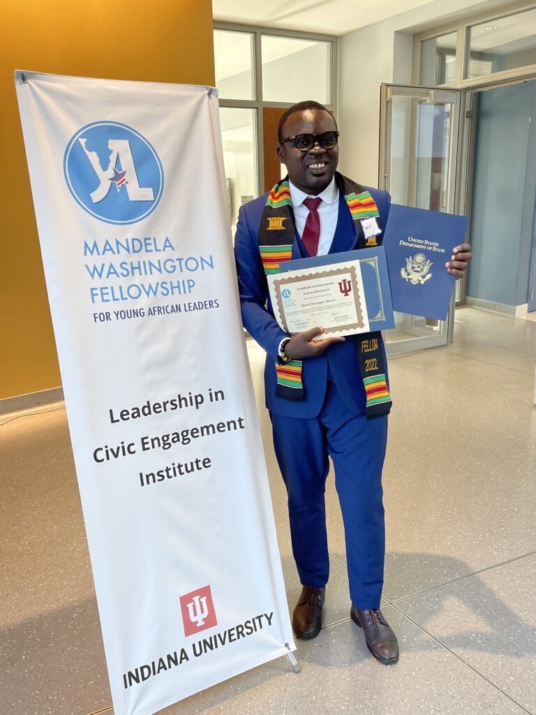 Daniel holding his graduation certificate in Leadership in Civic Engagement. He wears a blue suite, dark lens glasses, and a stole around his neck. To his right is a large banner that reads Mandela Washington Fellowship.