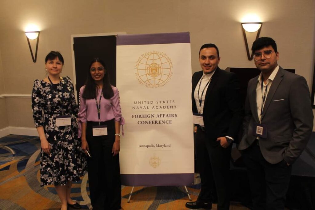 Dr. Mohan, wearing a gray suit jacket, stands next to other Fulbright Nehru scholars: 2 women and another man. Between them is a large banner that reads "Unite States Naval Academy Foreign Affairs Conference Annapolis, Maryland."