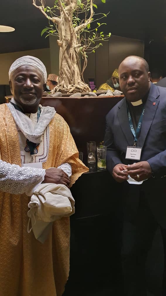 Reverend Mdumi in a blue pastor's suite standing to the right of Imaam Salih wearing traditional orange robes and partial white head covering, sometimes known as a turban or kufiya