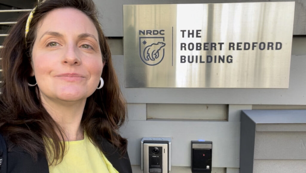 E 1 Hotel Fellowship winner, Mary Kombolias stands smiling in front of a plaque with the NRDC office logo and the words "The Robert Redford Building"