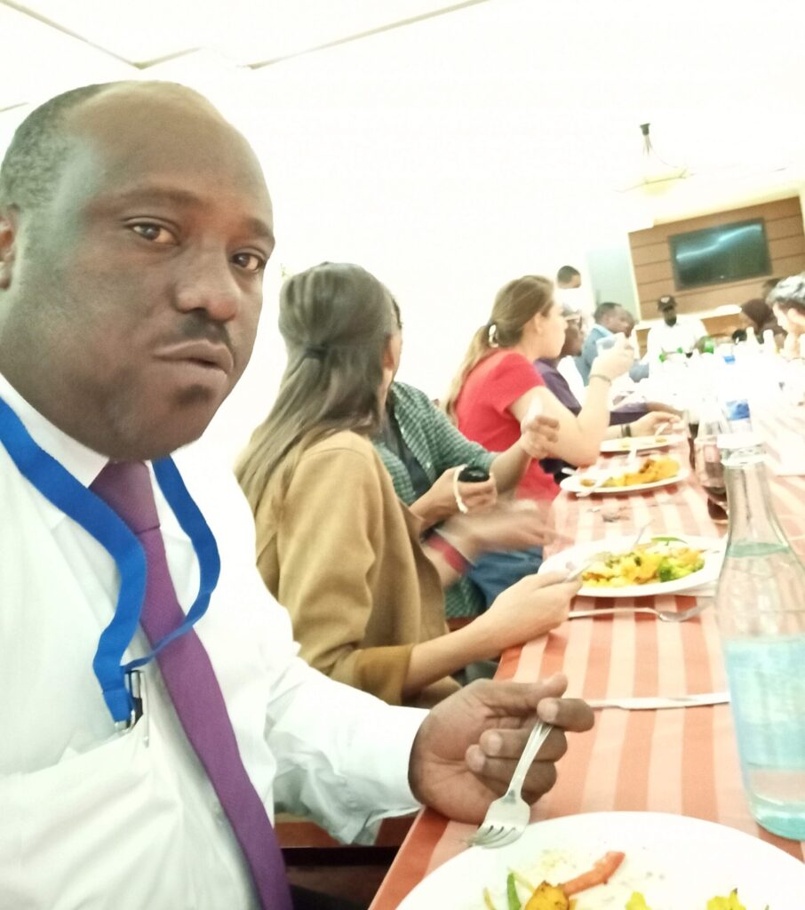 Rev. Mdumi wearing a white shirt, a maroon tie, and a blue lanyard around his neck. He sits at a banquet table, sharing a meal with other KAICIID Fellows.