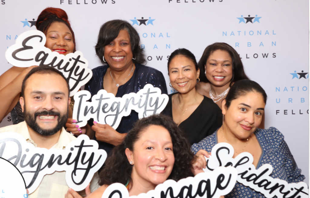 Six smiling NUF fellows pose with India Pierce Lee in front of a background with National Urban Fellows logos, holding up signs with the words: Equity, Integrity, Solidarity, and Courage