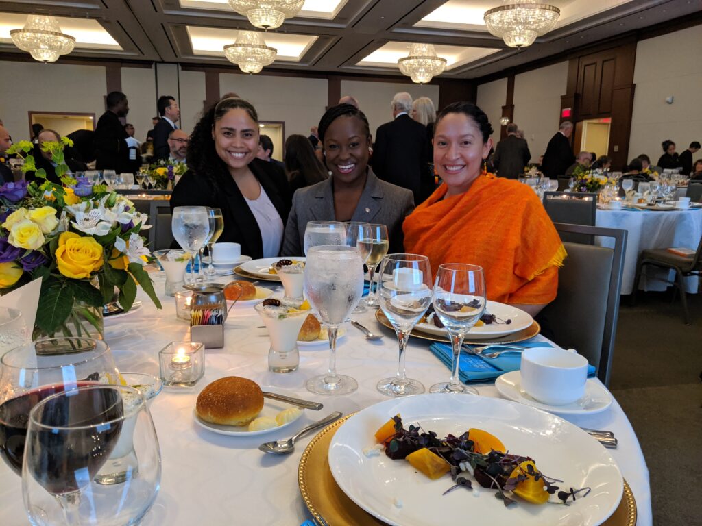 Angelique Salizan, Regina Johnson and Cari Morales attending a fundraiser during their time in Cleveland. National Urban Fellows Alumni Class of 2020. Fellows sit around a large circular table with food, glasses, and flower centerpieces.