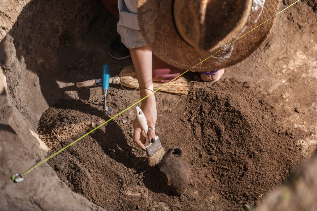 A female archaeologist wearing a wide-brimmed straw hat carefully excavates an antique ceramic object at an archaeological site.