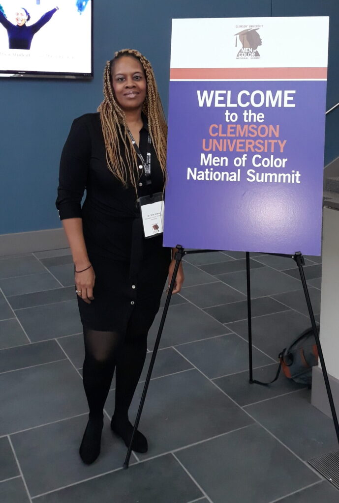 Dr. Nita Evans, dressed in formal black clothing, stands next to a purple and white sign reading "Welcome to the Clemson University Men of Color National Summit."