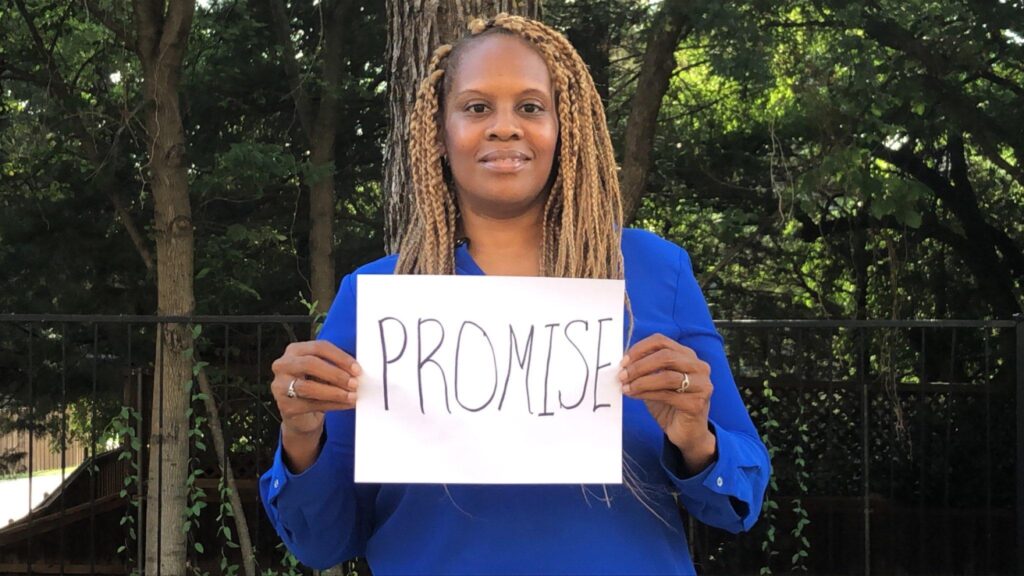 Nita Evans, who won the Mira Fellowship, in a blue shirt, smiles at the camera while holding a piece of paper reading "Promise."