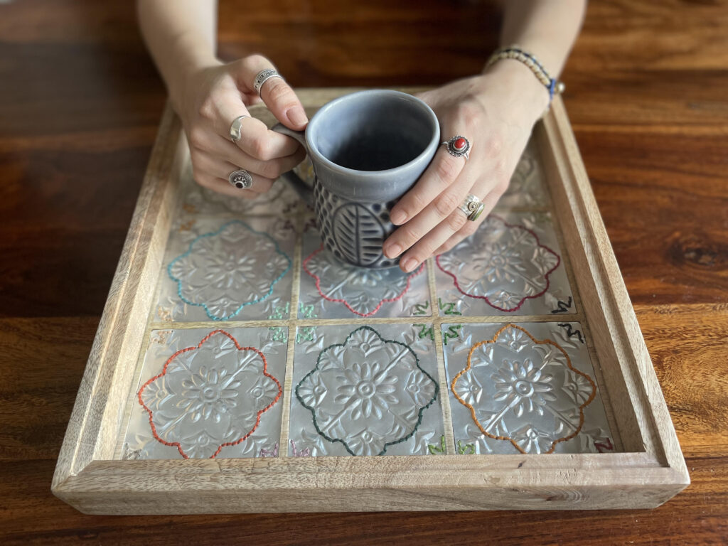 A pair of hands wearing several rings and bracelets holds a mug on top of an up-cycled tray created from cold beverage cans hand embossed by the project’s creative partners.