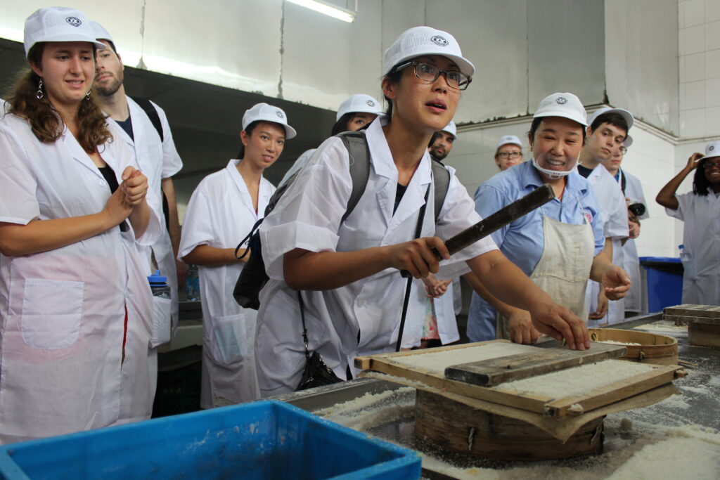 A student tries their hand at pounding some traditional sweets on a wooden block, fellow students all around them.