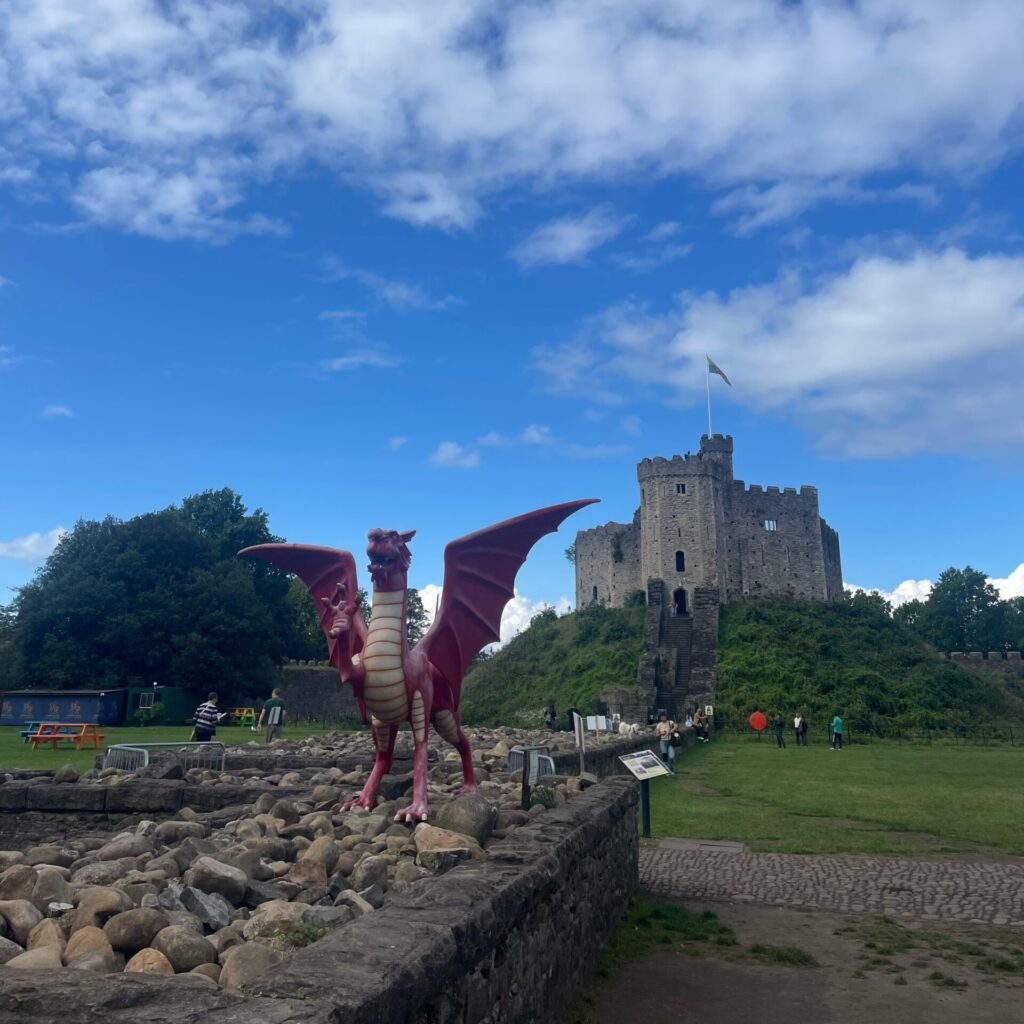 A statue of the red dragon stands in the foreground; in the background, Cardiff Castle, with the Welsh flag flying over it.