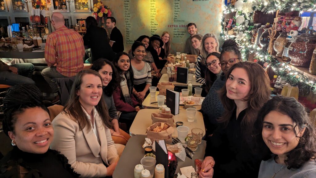 International Fellows Network (IFN) group sitting at tables arranged horizontally, smiling at the camera. Fellows sit on either side of the tables with appetizers on the tables and various decorations consisting of jars, holly, and lights that line the right-hand side wall.