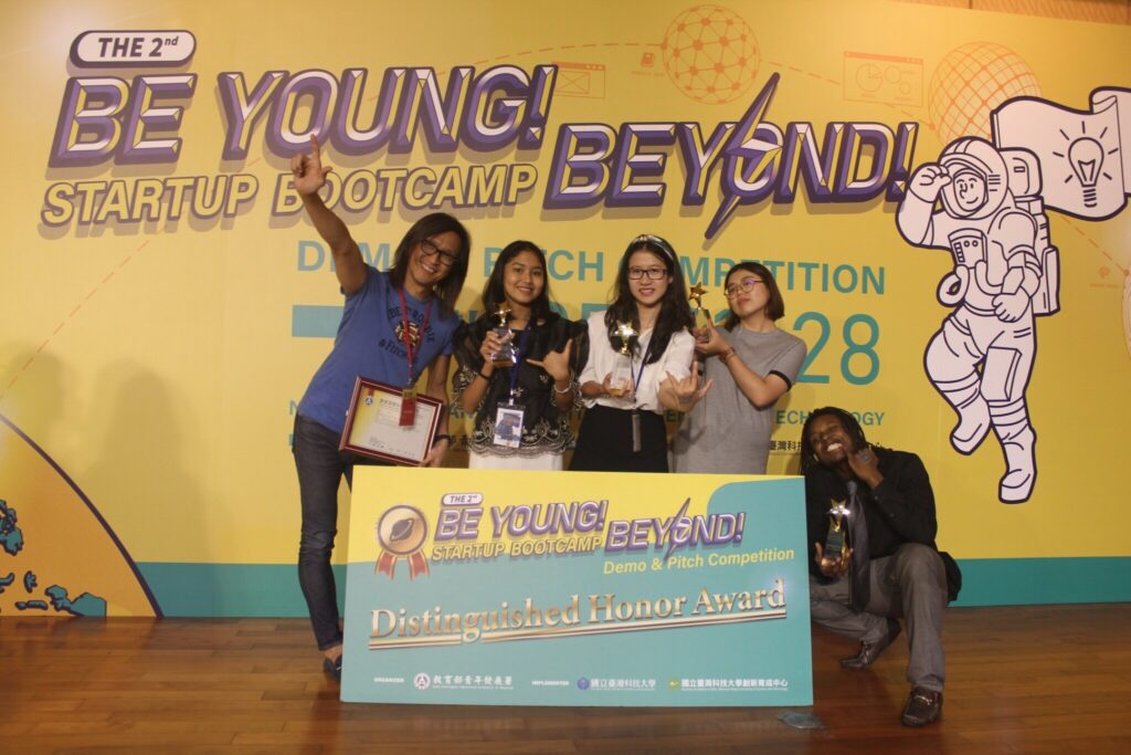 Miles, on the far right, and four other students from the Taiwan competition. The students are standing, smiling, and holding trophies. Miles is kneeling, wearing a black dress shirt and gray dress pants with a gray tie, smiling, and holding a trophy. The individuals are standing behind a small banner that reads "Be Young! Beyond! Startup Pitch Competition". A large poster behind them reads the same message.
