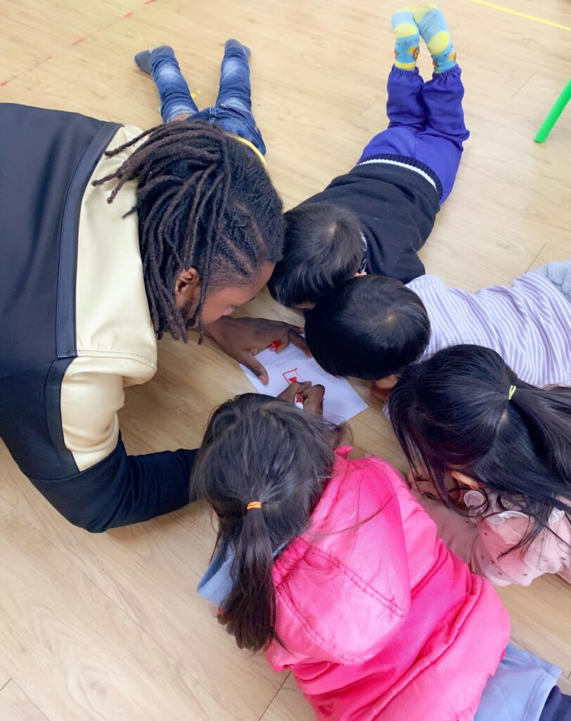 Miles Iton, in Taiwan laying on the floor with preschool students around him, as he writes on a piece of paper with an orange marker.