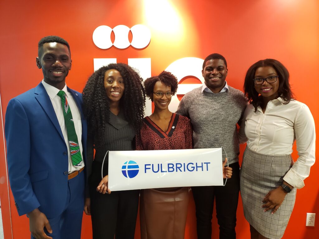 Olugbenga, in a blue suit on the left, stands with a group of other Fulbright recipients. One in the center is holding a sign with the Fulbright logo. The wall behind them is orange.