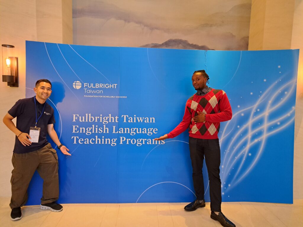 Fulbright Awardee Miles Iton standing in front of a large blue poster that reads Fulbright Taiwan English Language Teaching Program. Miles is wearing a red, diamond-patterned sweater with black dress pants and shoes and points to the words on the poster. A Staff member on the left-hand side does similarly.