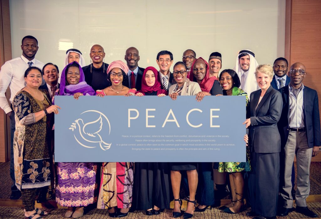 A large group of diverse people from all over the globe holding a large blue placard that says peace with a vector image of a white dove holding an olive branch.