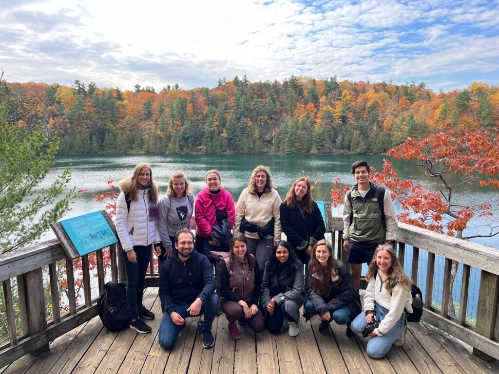 Fulbright Killam Fellow Anusha Natarajan taking a group photo with other fellows at a park in Quebec. The Fellows don jackets and are stand on a wooden viewing area that overlooks a lake with fall trees in the background.