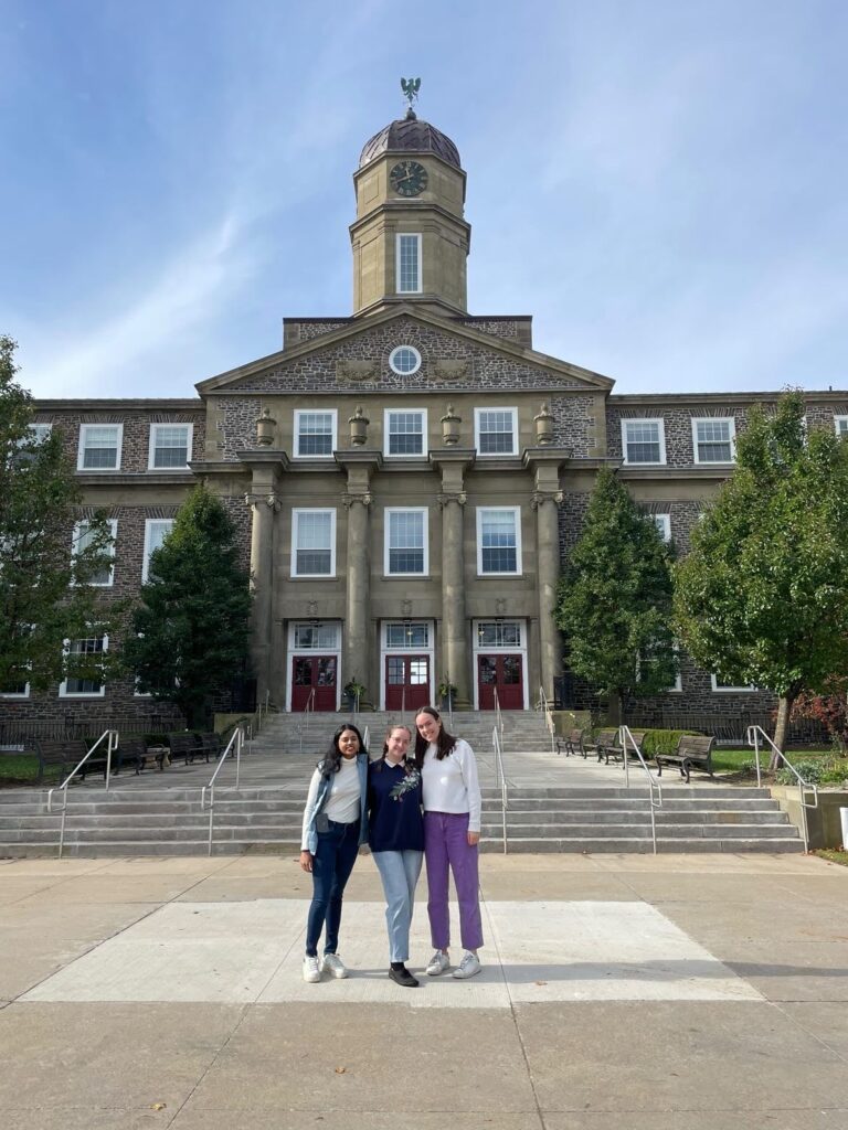 Fulbright Killam Fellow Anusha Natarajan with two friends standing in front of a large university building with a clocktower vestibule, in front of stairs. Dark green trees are at the edges of the photo.
