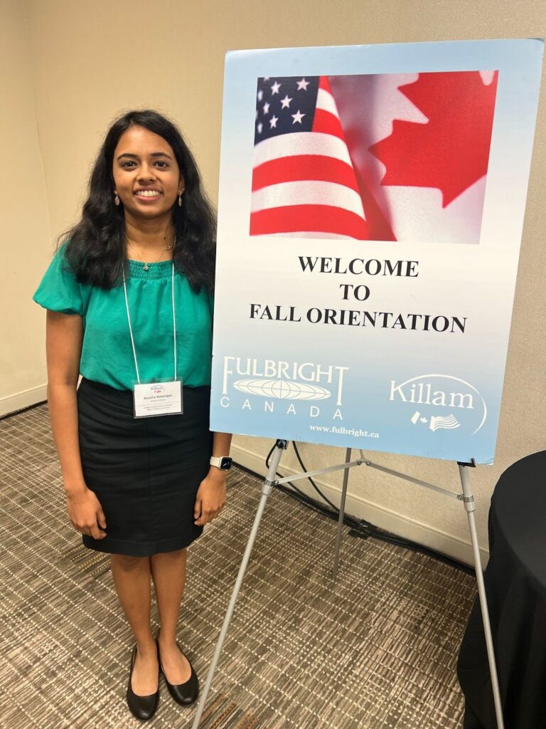 Fulbright Killam Fellow Anusha Natarajan wearing business attire with a teal green top and black skirt. She stands next to a sign that reads "Welcome to Fall Orientation"