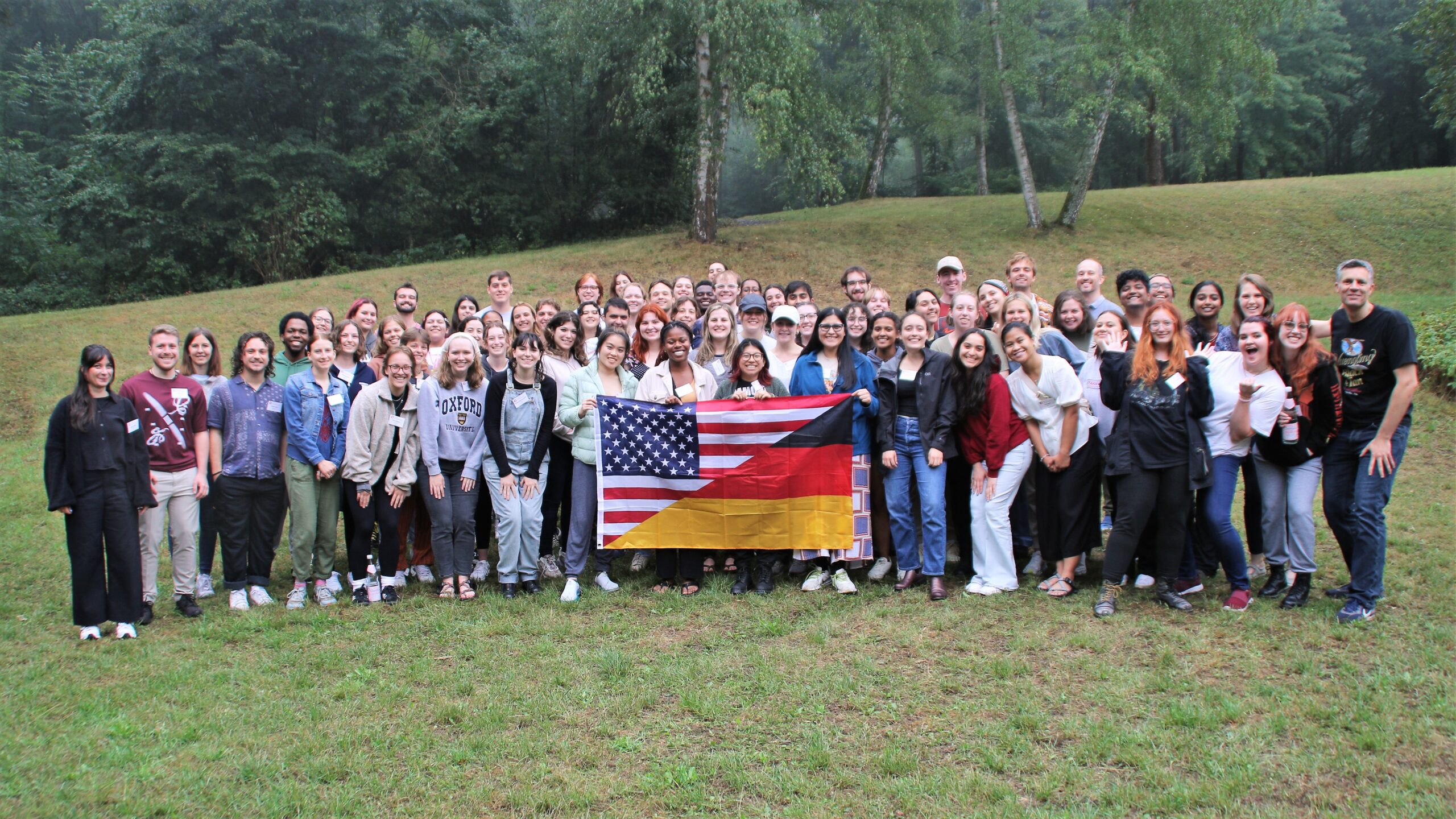 The 40th CBYX cohort poses for a large group photo in an open field with trees in the background. In the middle, they hold a flag which is a diagonal split between the flags of the US and Germany.