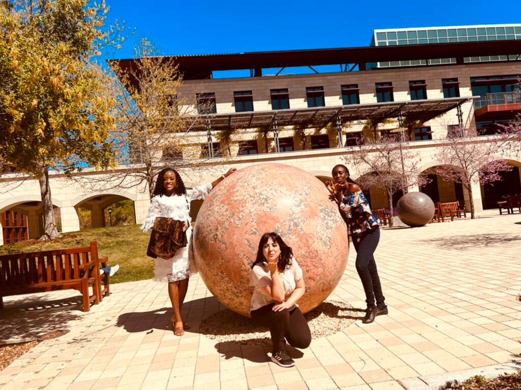 Fellowship winner Amanda Obidike wearing a white dress standing next to a spherical art installation at Sandford, along with 2 other TechWomen fellows at the university's campus.