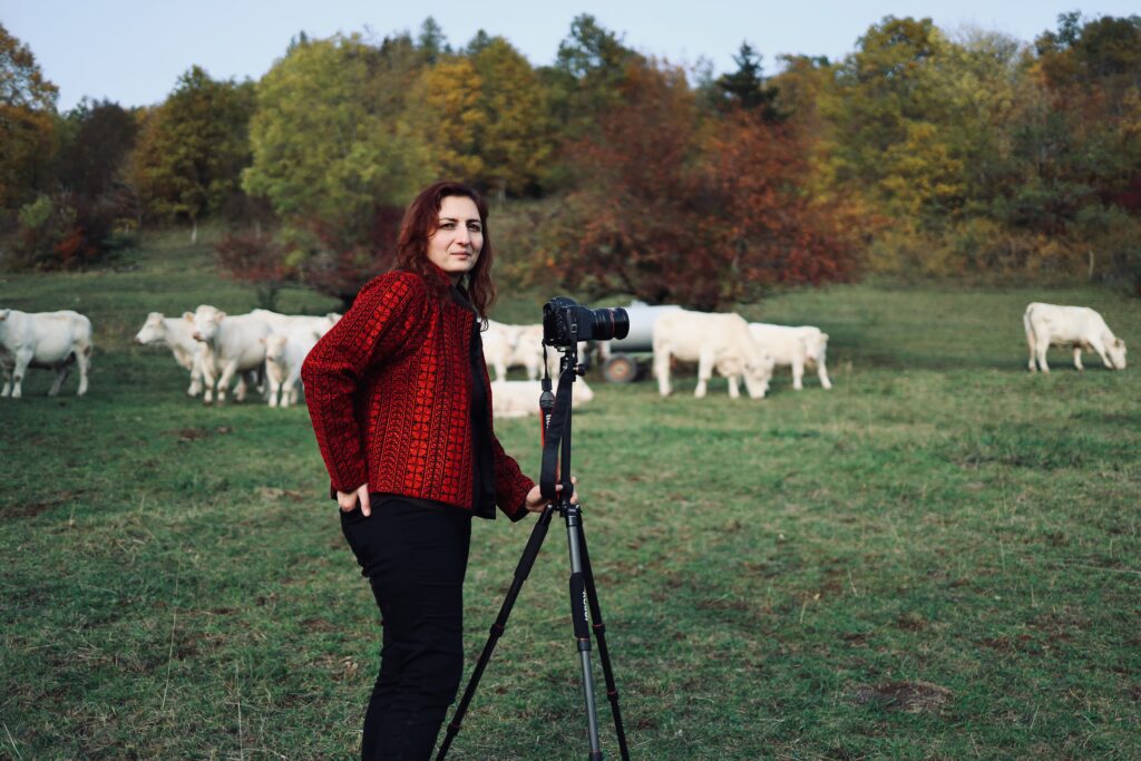 Dr. Tibet, wearing black pants and a red jacket, stands in a green field with a camera mounted on a tripod. Behind her stand cows and trees.