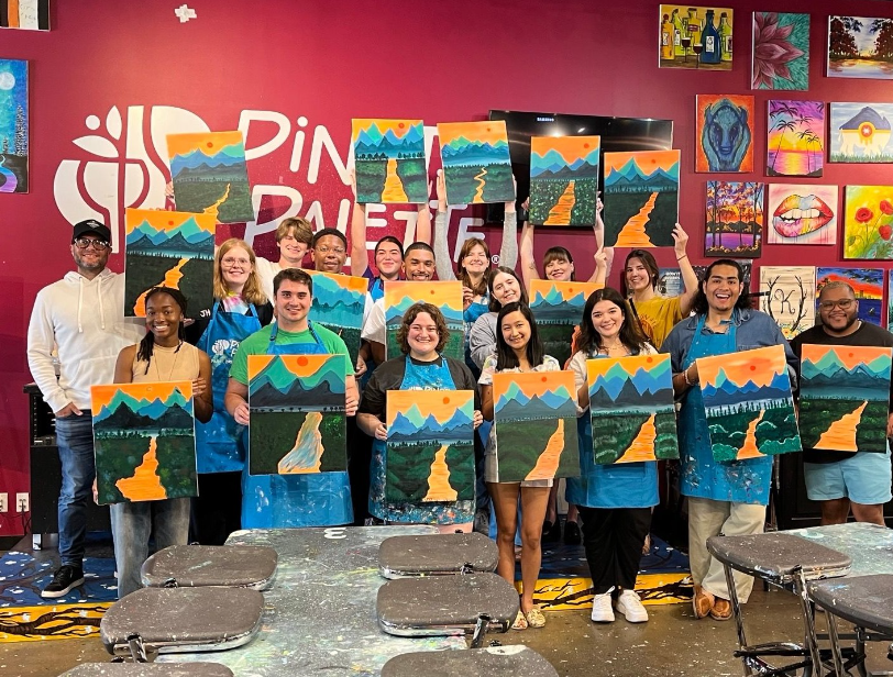 The Tulsa Service Year cohort hold up a series of similar paintings depicting a river, mountains, and the sun. Behind them is a red wall reading "Pinot's Palette" with other paintings on the wall.