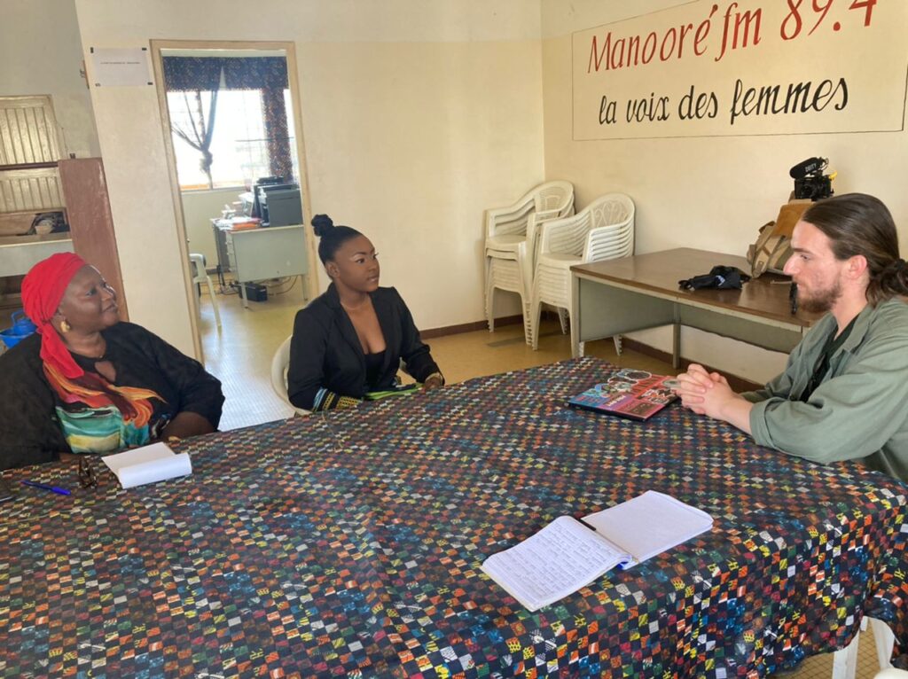 Gilman and McNair Scholar Rowan Glass sitting at a table with brightly colored and checkered tablecloth, with Manooré FM radio staff members.