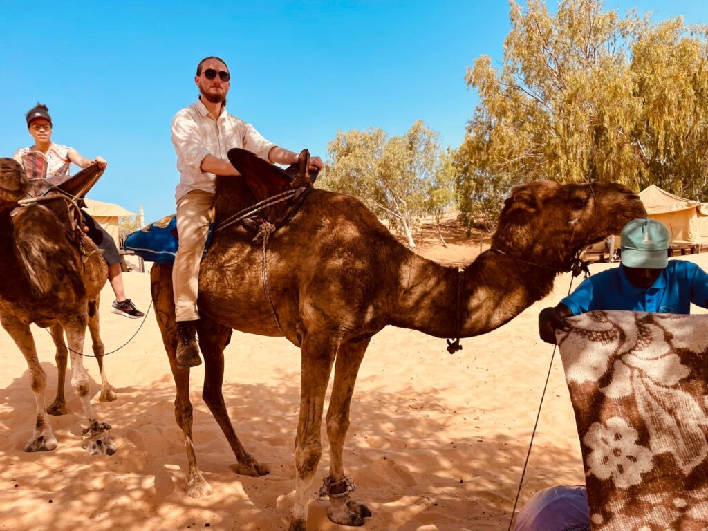 Rowan Glass, Gilman Scholar, wearing a white short and khaki pants, riding a brown camel in the desert.