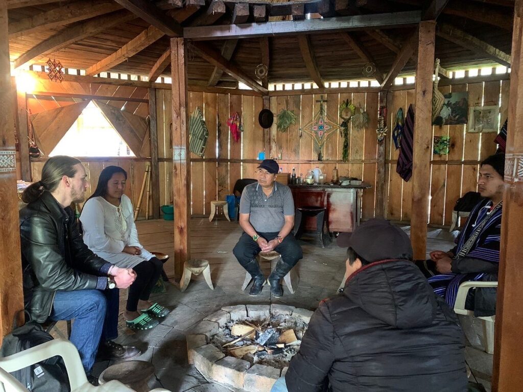 Gilman and McNair Scholar Rowan Glass sitting inside a wooden roundhouse, around a fire pit. Other people sit in plastic chairs around the fire put as well. Many artifacts are hung up on the walls of the roundhouse.