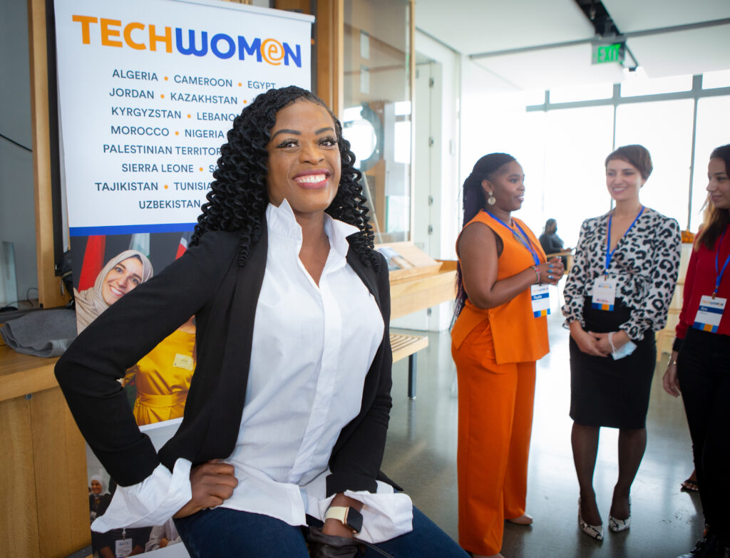 Amanda Obidike, TechWomen Fellowship winner, smiling happily at the caerma, wearing a white dress shirt and black suite jacket. She's sitting, one hand on her side, slighting leaning forward in front of a TechWomen banner.