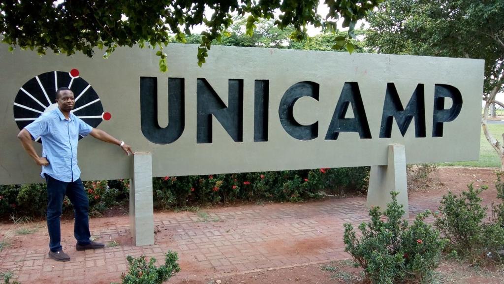 Fiocruz Scholar, Chinedu Okeke, at the main entrance of The State University of Campinas, in Campinas, Brazil. He wears a blue button-up shirt while resting his hands on the large University sign that reads "UNICAMP."