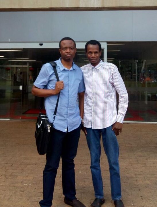Chinedu, wearing a blue button up collard shirt, with a bag over his shoulder, standing next to his friend from Nigeria. The partial name of the library can be seen behind the men.