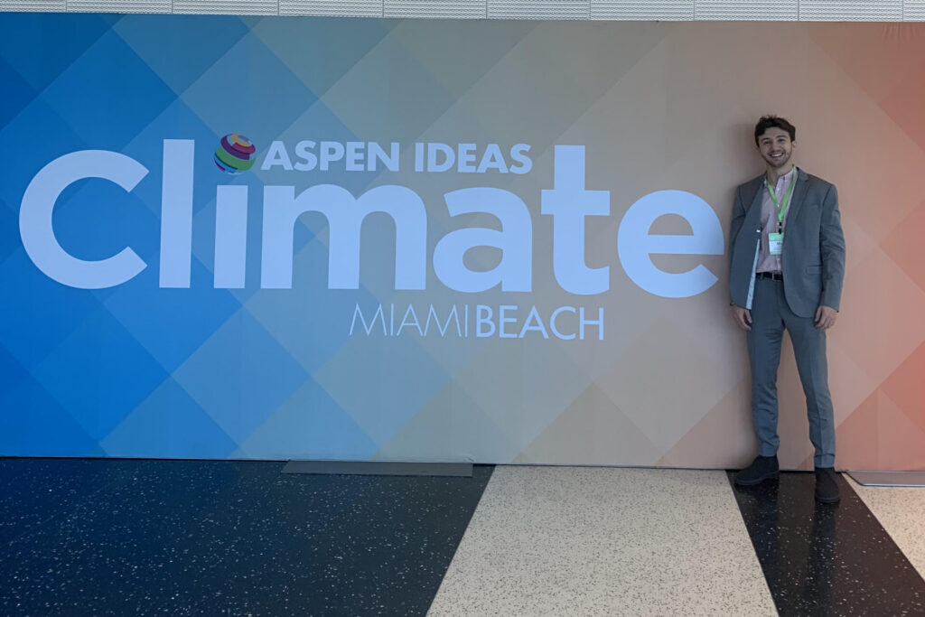Max, wearing a formal gray suit, stands to the side of a large poster on a wall which reads "Aspen Ideas Climate Miami Beach."