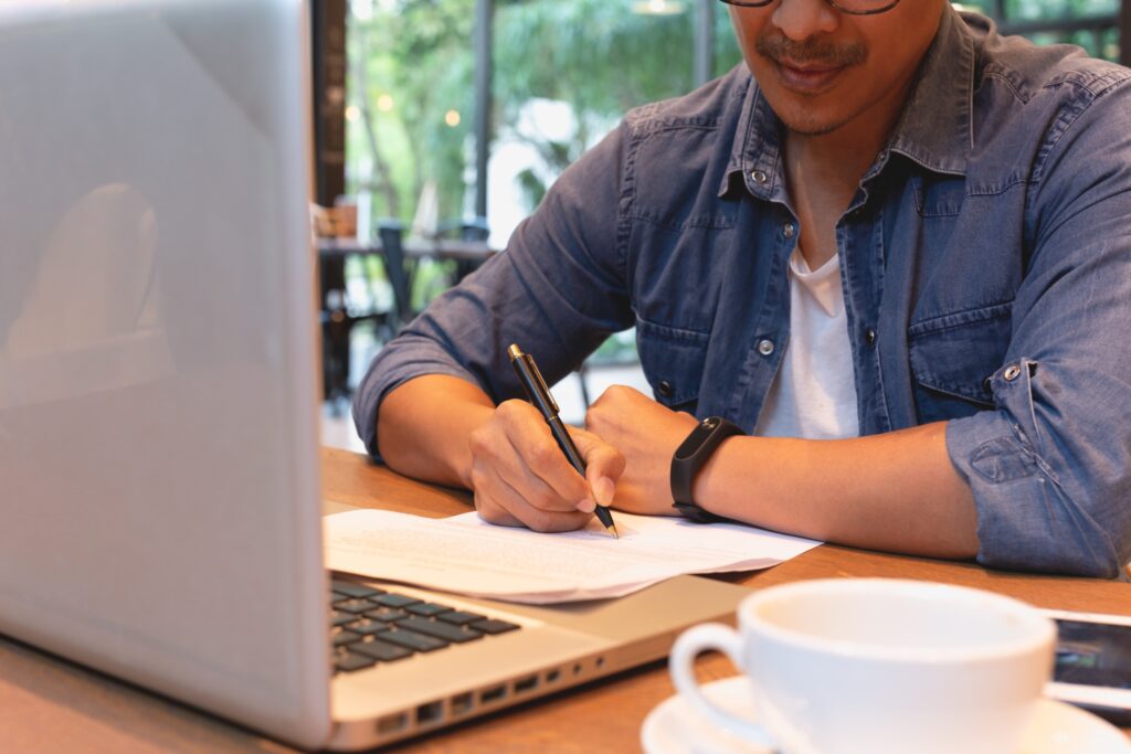 A young man sitting at a coffee shop, with a laptop and a coffee cup. He is writing on a piece of paper with a pen. It is representative of the upcoming fellowship deadlines for the Emerging Writer Fellowships.