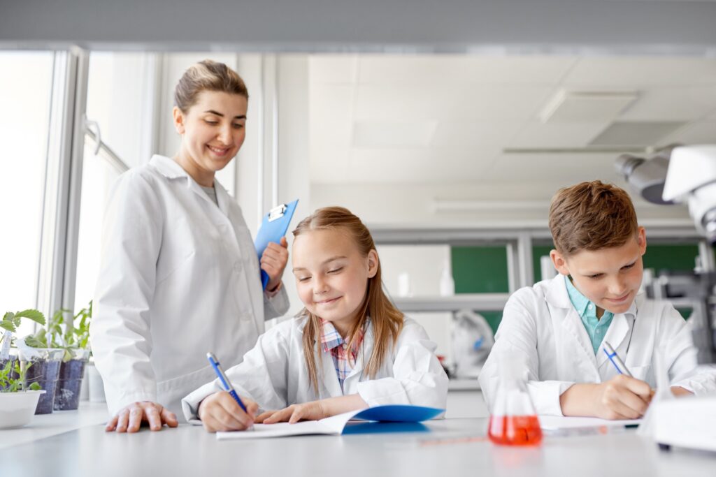 Young science teacher, wearing a lab coat, teaching 2 elementary school students sitting at their workbench, writing in their notebooks. The students, a girl and a boy, sit next to each other and are also wearing lab coats. There is an Erlenmeyer flask filled with red liquid and a microscope partially outside of the frame. The image represents the upcoming fellowship deadline for a STEM teacher fellowship.