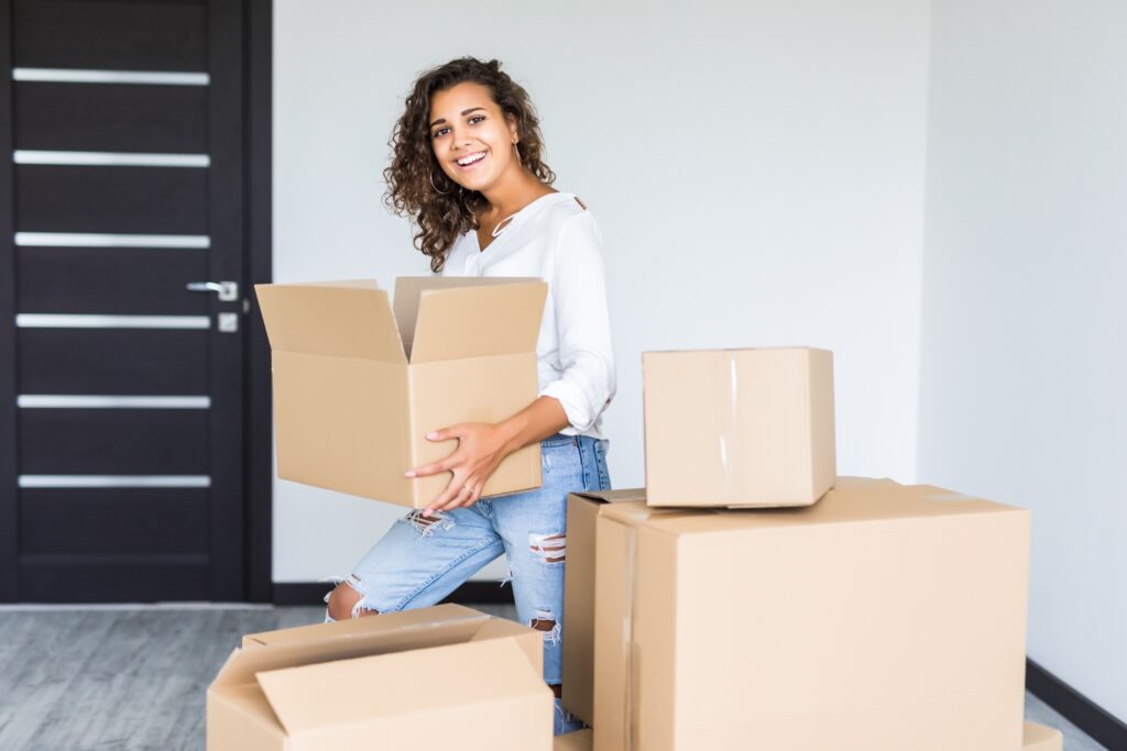 Young ethnically diverse woman with curly hair, holding a moving box smiling at the camera. She is in an empty apartment, with a black door, and there are moving boxes around her. The image signifies tips for moving abroad for an international fellowship.