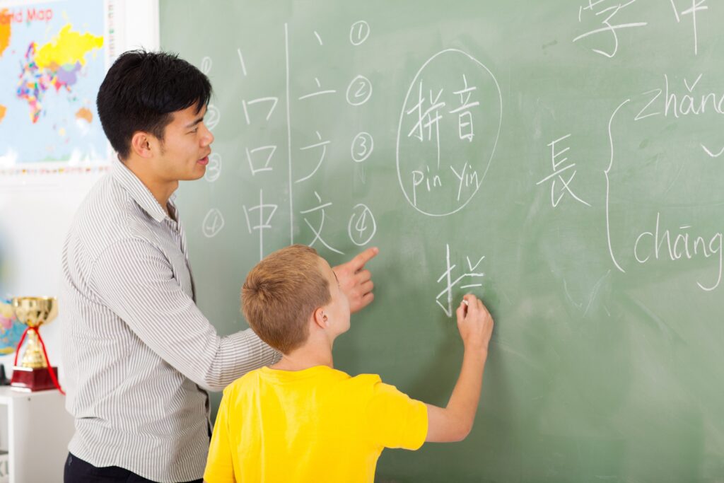 Young Asian man wearing a stiped dress shirt pointing to blackboard, teaching Chinese to an elementary school student wearing a yellow shirt, writing a Chinese character with chalk. This image is representative of the upcoming fellowships deadline for the Chinese Language Program.