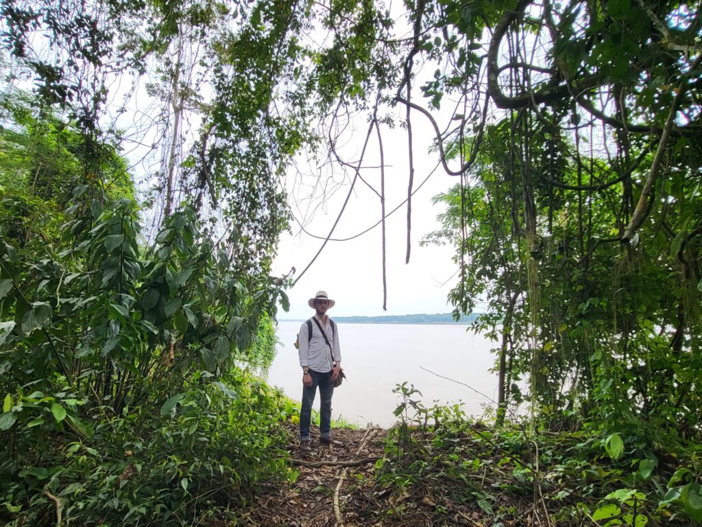 Multi fellowship winner, Rowan Glass, wearing a panama hat and a white button-up shirt, stands in a jungle clearing with the Amazon River visible in the background.