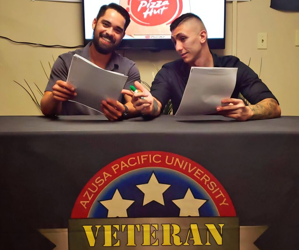 Angel Reyes, right, sitting next to friend David Eskridge sitting at at a table with the label "Azusa Pacific University, Veteran," an even that was made possible by the mentorship from his mentor Blason Taon, not pictured.