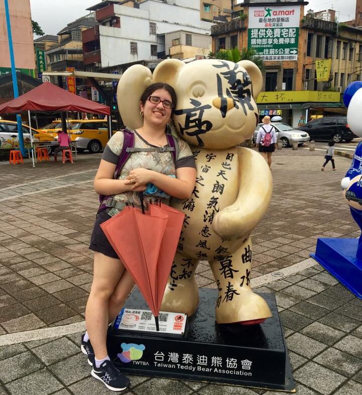 Fulbright winner Gina Elia, wearing a t-shirt and shorts and holding a red umbrella, stands next to a bear statue with Chinese characters in the midst of an urban courtyard.