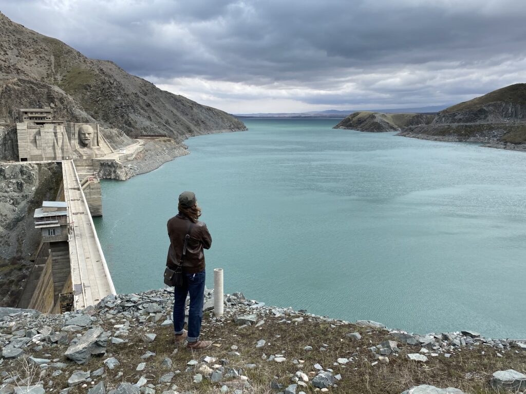Rowan Glass, a young man, winner of multiple fellowships, wearing a leather jacket, green baseball cap, jeans, and leather boots, stands on a cliffside overlooking a large reservoir in the mountains of Kyrgyzstan, during his international off the beaten path travel.