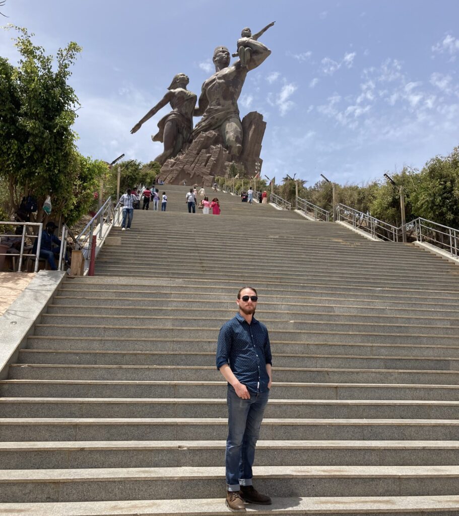 Rowan Glass, wearing a blue button-up shirt and jeans, stands on a large stone staircase leading to the towering African Renaissance Statue in Dakar, Senegal, during his fellowship program.