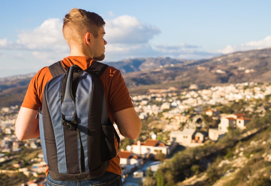 Young man wearing a blue and black backpack, his back turned to the camera, looking over a town at the end of a hill. He represents a student taking a gap year to travel.