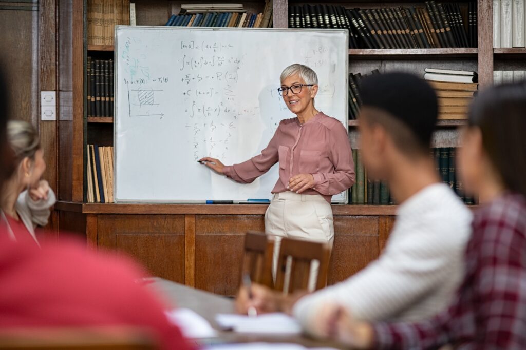 Senior teacher explaining math formulas written on whiteboard in library while college students sitting on table understanding the concept. Happy mature woman lecturer clearing doubts to students in class. Professor teaching to high school guys and girls. The image is representative of a Fulbright Scholar winner.